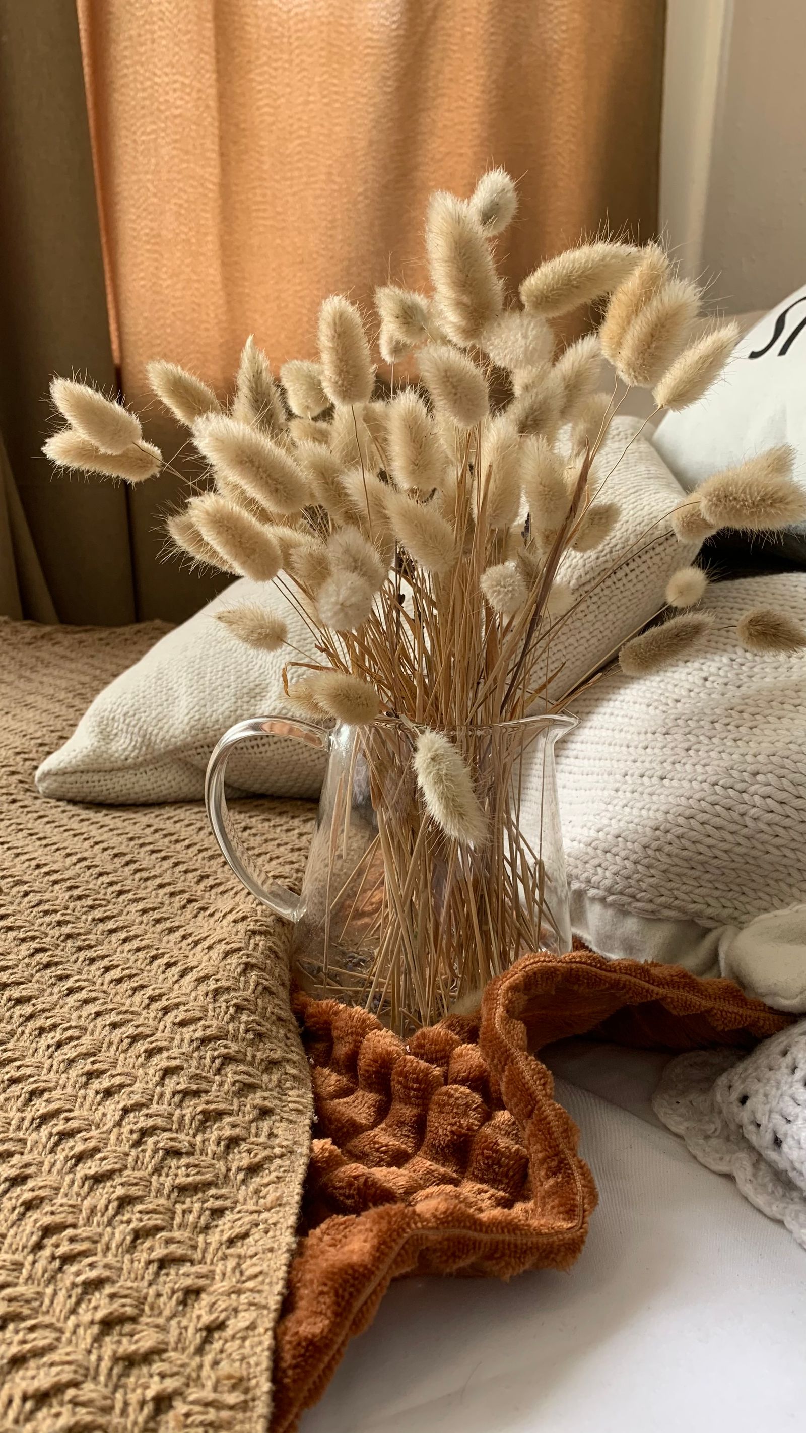Dried bunny tail grass in a glass pitcher, with a rust textile and a cream knit throw