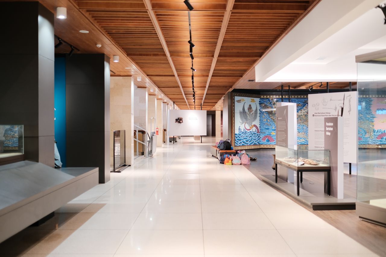 Museum exhibition gallery with a timber-slat ceiling, track lighting, freestanding display cases and a painted mural